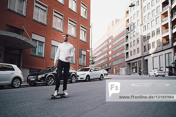 Junger Mann fährt in voller Länge mit dem Skateboard auf der Straße gegen Gebäude in der Stadt