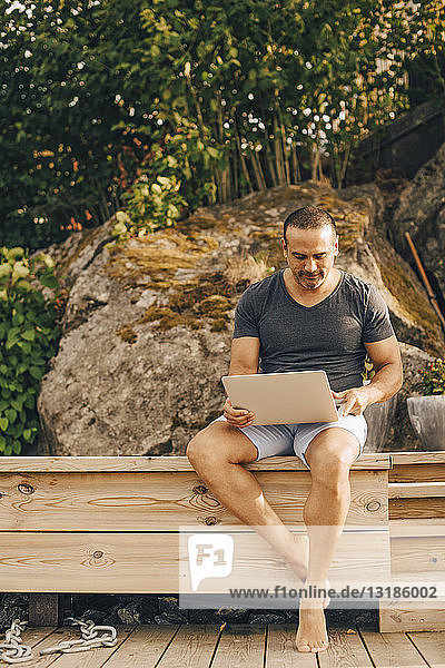Erwachsener Mann in voller Länge mit Laptop  während er auf der Terrasse gegen Bäume sitzt