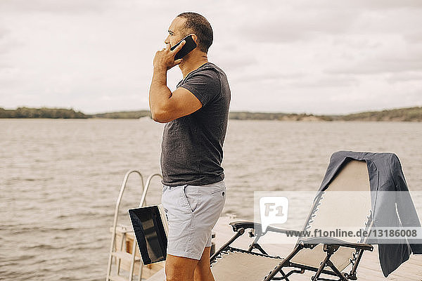 Mann mit Laptop spricht mit einem Smartphone  während er auf einem Steg am See gegen den Himmel steht
