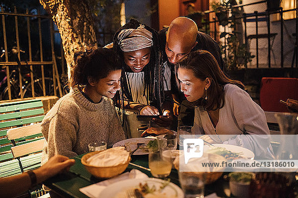 Glückliche junge Frau zeigt Freunden bei Tisch während der Dinnerparty ihr Smartphone