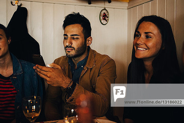 Junger Mann benutzt Smartphone  während er während einer Dinnerparty im Restaurant inmitten von Freundinnen sitzt