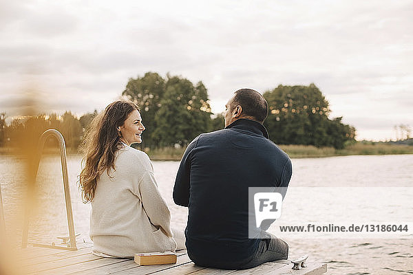 Smiling couple talking while sitting on jetty by lake against sky