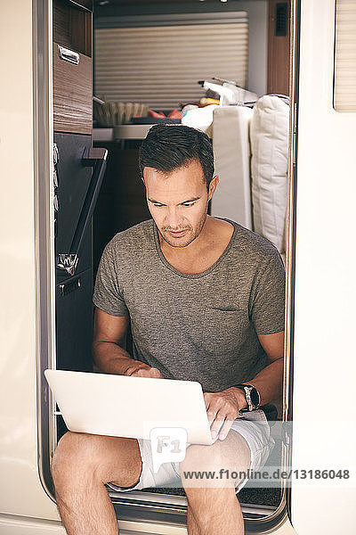 Reifer Mann benutzt Laptop  während er auf der Tür eines Wohnwagens sitzt