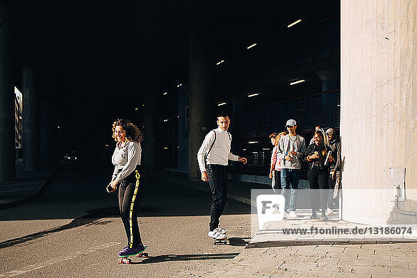 Freunde schauen Mann und Frau beim Skateboarden auf der Straße an einem sonnigen Tag zu