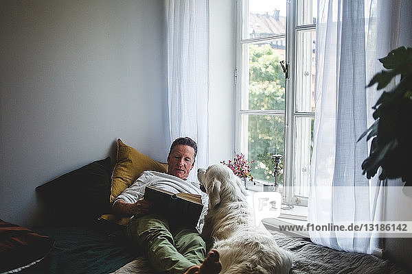 Älterer Mann liegt mit Hund  während er zu Hause auf dem Bett am Fenster ein Buch liest