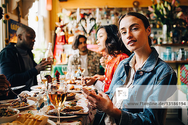 Porträt einer selbstbewussten jungen Frau  die während der Dinnerparty mit einem Smartphone gegen Freunde am Tisch im Restaurant sitzt