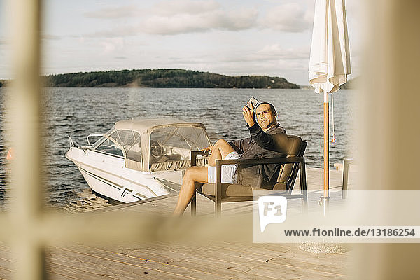 Ein erwachsener Mann  der auf der Terrasse am See sitzt und sich mit seinem Handy selbstständig macht