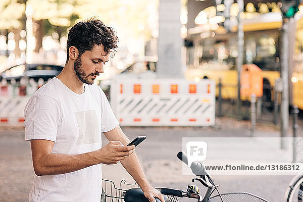 Junger Mann benutzt Mobiltelefon  während er mit dem Fahrrad auf der Straße in der Stadt steht