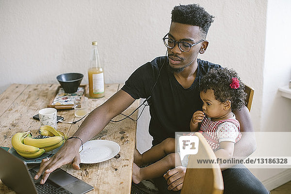 Junger Mann benutzt Laptop  während er mit seiner Tochter am Esstisch im Haus sitzt