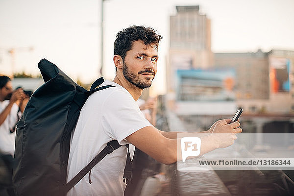 Porträt eines jungen Mannes in Seitenansicht  der ein Mobiltelefon in der Hand hält  während er sich an der Brücke auf ein Geländer stützt