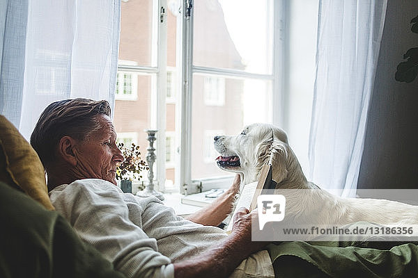 Älterer Mann liest Buch  während er sich zu Hause mit Hund im Bett entspannt