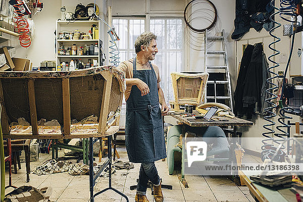 Mature male craftsperson looking away while standing by incomplete sofa in upholstery workshop