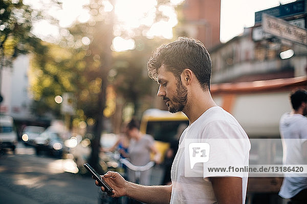 Seitenansicht eines jungen Mannes  der ein Mobiltelefon benutzt  während er in der Stadt auf der Straße steht