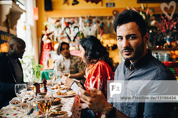 Porträt eines selbstbewussten jungen Mannes  der ein Smartphone in der Hand hält  während er mit Freunden im Restaurant während des Dinner-Partes am Tisch sitzt