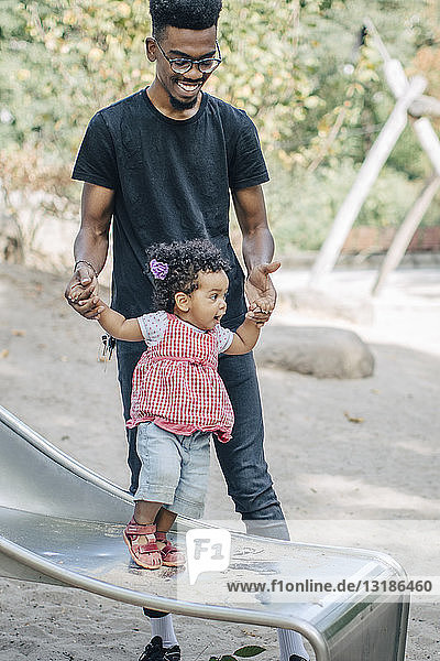 Junger Mann hält Hände einer Tochter  die auf einer Rutsche auf einem Spielplatz steht