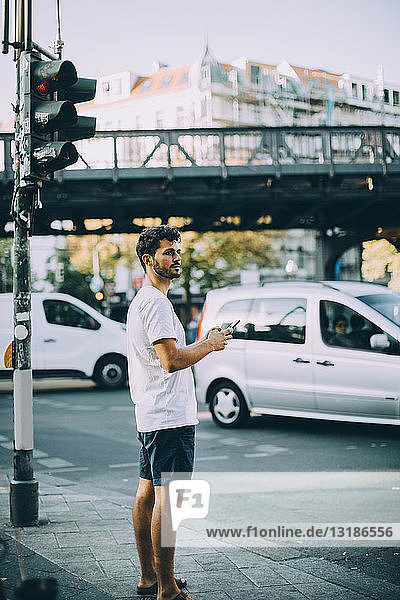Side view of young man holding mobile phone while standing on sidewalk in city