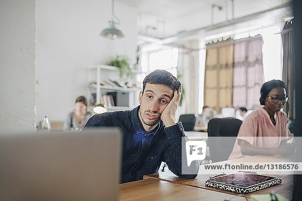 Müde Geschäftsmann schaut auf den Laptop  während er am Schreibtisch im Kreativbüro sitzt