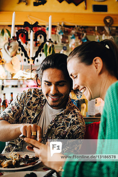 Smiling young man pointing at smart phone to woman sitting in restaurant during brunch party
