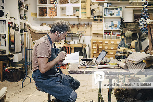 Seitenansicht eines männlichen Polsterers  der an der Werkbank in der Werkstatt sitzt und ein Buch liest