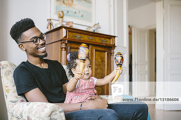Kleines Mädchen hält Maracas  während es zu Hause mit dem glücklichen Vater auf einem Sessel sitzt