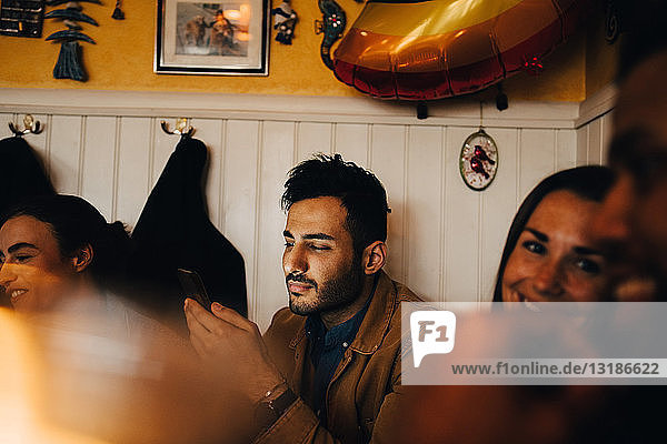 Junger Mann benutzt Mobiltelefon  während er inmitten von Freunden im Restaurant sitzt