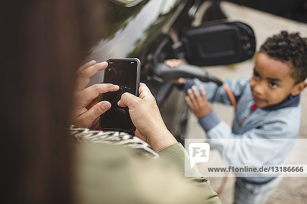 Hochwinkelansicht eines Jungen  der ein Elektroauto auflädt  während er die Mutter mit einem Smartphone anschaut