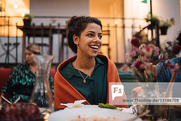 Lächelnde junge Frau schaut weg  während sie während der Dinnerparty am Tisch sitzt