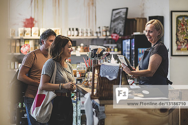 Smiling female sales clerk looking at food product while customers standing at checkout counter