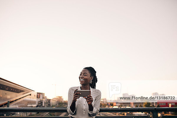 Smiling young woman looking away while holding mobile phone against clear sky in city