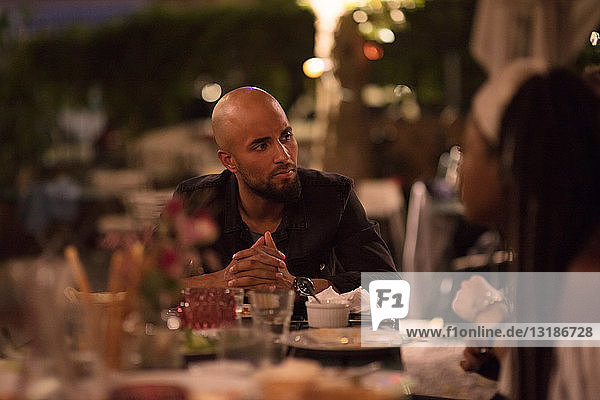 Young man with shaved head looking at female friend while sitting at table during dinner party