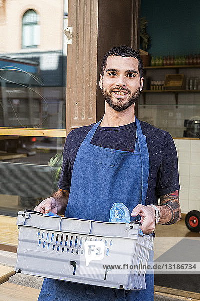 Porträt eines lächelnden jungen männlichen Besitzers  der eine Kiste gegen ein Restaurant trägt