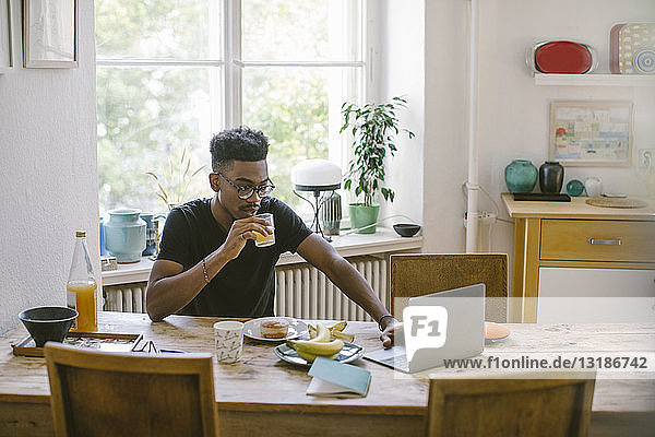 Junger Mann trinkt Saft  während er am Tisch im Haus auf seinen Laptop schaut