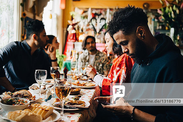 Junger Mann verschickt Textnachrichten über Smartphone  während er mit Freunden während einer Brunch-Party im Restaurant sitzt