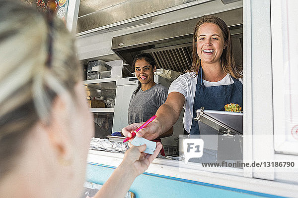 Smiling young female owner standing by colleague in food truck giving paper to customer