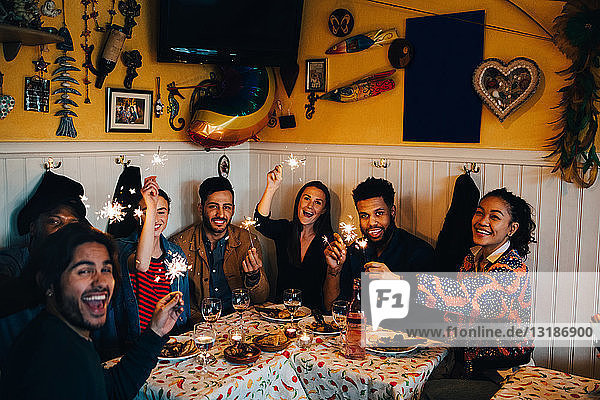 Porträt fröhlicher junger multi-ethnischer Freunde  die mit Wunderkerzen am Tisch im Restaurant sitzen