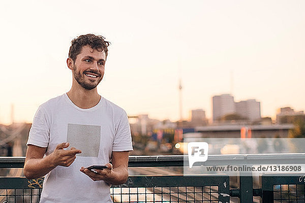 Lächelnder junger Mann schaut weg  während er bei Sonnenuntergang sein Handy auf der Brücke gegen den klaren Himmel hält