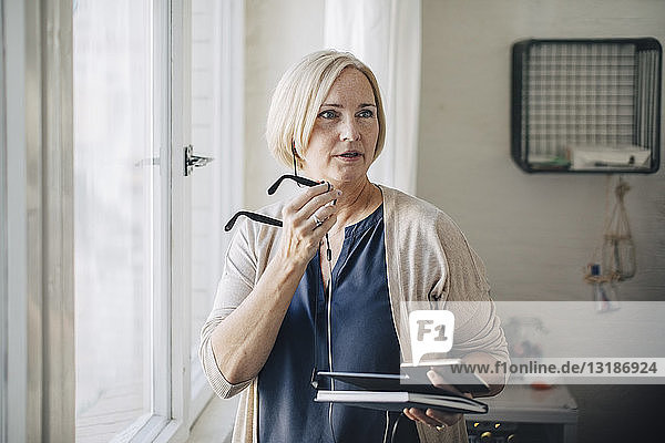 Mature businesswoman talking through earphones while standing by window in office