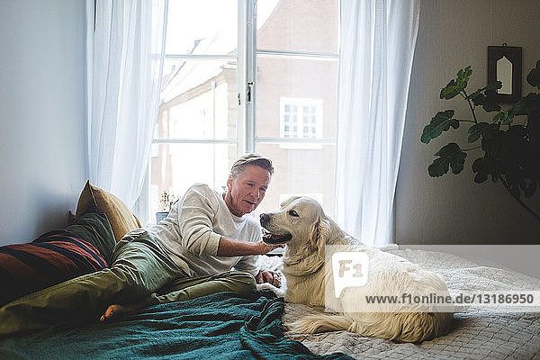 Senior man stroking dog while leaning on bed at home