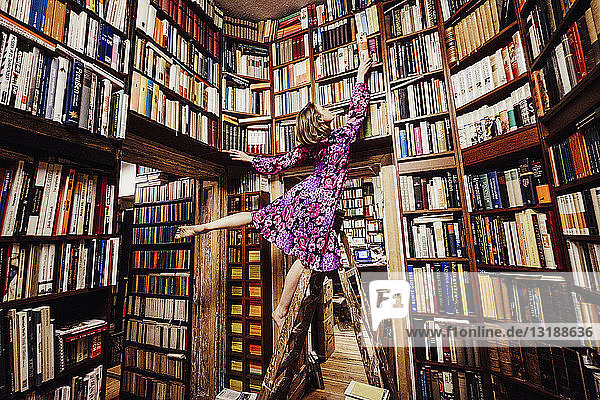 Unbekümmerte Frau auf einer Leiter  die nach einem Buch in einer Bibliothek greift