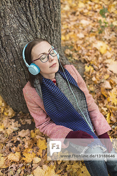 Serene woman listening to music with headphones below tree in autumn park