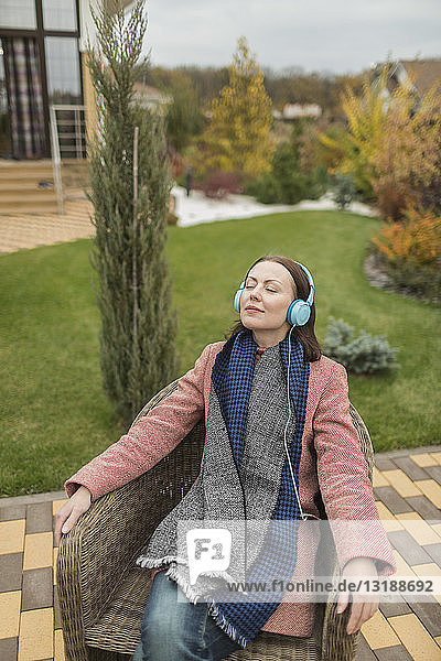 Serene woman in scarf and coat listening to music with headphones on autumn patio