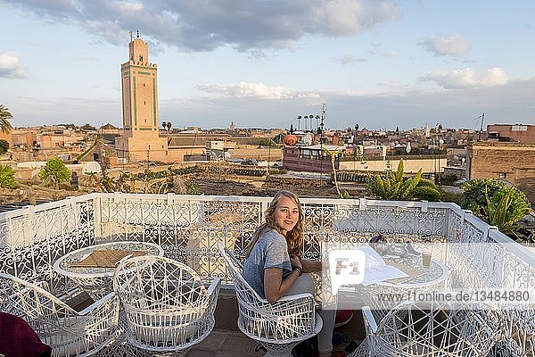 Junge Frau auf einer Dachterrasse liest die Speisekarte im Restaurant  Blick auf die Altstadt  Moschee mit Minarett  Abendstimmung  Marrakesch  Marokko  Afrika