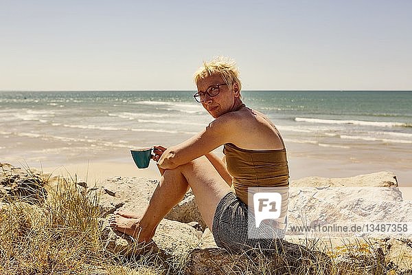 Frau  sitzt auf einem Felsen am Meer und trinkt eine Tasse Kaffee  Camping  Portbail  Normandie  Frankreich  Europa