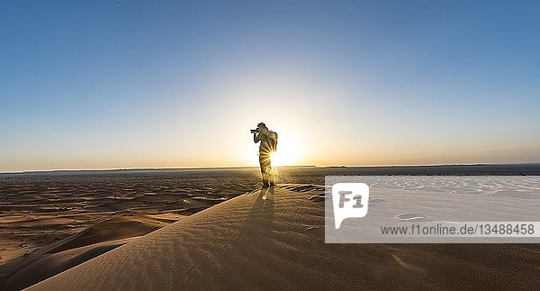 Young man standing on sand dune and photographed  sunbeams at sunrise  Erg Chebbi  Merzouga  Sahara  Morocco  Africa