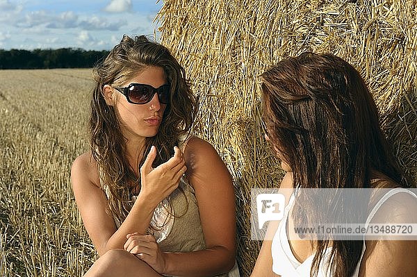 Zwei junge Frauen sitzen auf einem abgeernteten Getreidefeld vor einem Strohballen und unterhalten sich  Departement Finistere  Region Bretagne  Frankreich  Europa