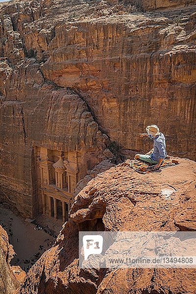 Tourist mit Sonnenhut sitzt auf Felsen und schaut von oben in die Schlucht Siq  in den Fels geschlagenes Schatzhaus des Pharaos  Fassade des Schatzhauses Al-Khazneh  Khazne Faraun  Mausoleum in der Nabatäerstadt Petra  nahe Wadi Musa  Jordanien  Asien