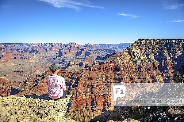 Junge Frau schaut in die Ferne  am Abgrund der gigantischen Schlucht des Grand Canyon sitzend  Blick vom Rim Trail  zwischen Mather Point und Yavapai Point  erodierte Felslandschaft  South Rim  Grand Canyon National Park  bei Tusayan  Arizona  USA  Nordamerika