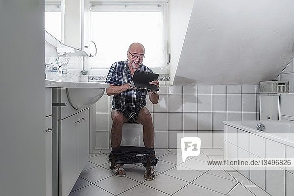 Senior man sitting on the toilet  laughing whilst surfing the internet on a tablet  Germany  Europe
