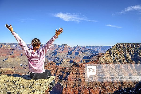 Junge Frau  die mit ausgestreckten Armen glücklich in die Ferne schaut  sitzend am Abgrund der gigantischen Schlucht des Grand Canyon  Blick vom Rim Trail  zwischen Mather Point und Yavapai Point  erodierte Felslandschaft  South Rim  bei Tusayan  Arizona  USA  Nordamerika