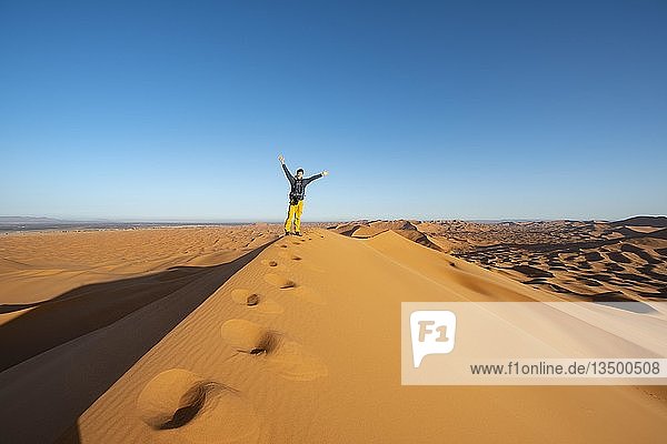 Junger Mann auf Sanddüne streckt seine Arme in die Luft  Erg Chebbi  Merzouga  Sahara  Marokko  Afrika